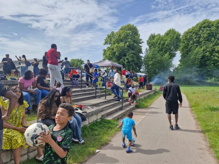 Children and families on the terrace steps beside the pitch, one child carrying a football, at the ZNVA Refugee Week family fun day, June 2026