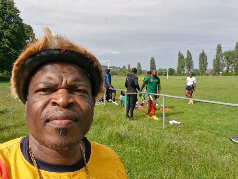 A ZNVA volunteer in a yellow shirt at the Refugee Week football tournament, with play continuing behind, June 2026
